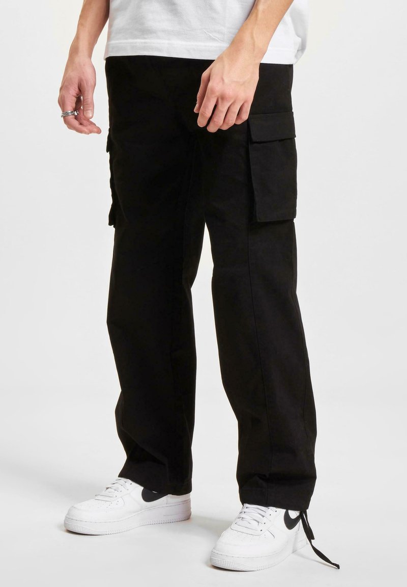 Black cargo pants with multiple side pockets and a relaxed fit, made of a sturdy fabric, worn with white sneakers.