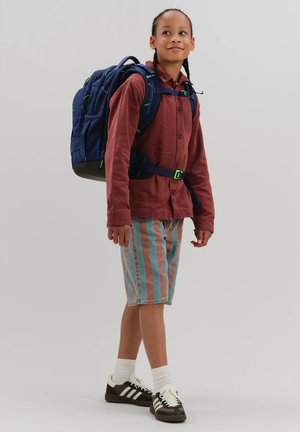 Child with braided hair wearing a red jacket, striped shorts, white socks, black and white shoes, carrying a large blue backpack.