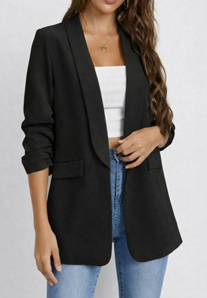Femme portant un blazer noir ouvert sur un top court blanc et un jean bleu, aux longs cheveux ondulés et portant un collier en or.