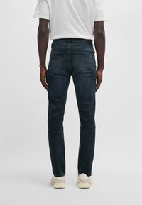 Dark blue denim jeans with a slim fit. Features a mid-rise waist, two back pockets, and a subtle faded texture. Styled with a plain white t-shirt.