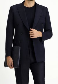 Granatowa dwurzędowa marynarka z wyłogami typu peak, w zestawieniu z czarną koszulą i spodniami. Trzyma czarną, fakturowaną kopertówkę z zamkiem błyskawicznym.