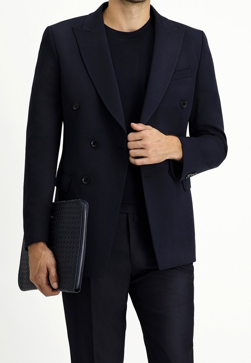 Granatowa dwurzędowa marynarka z wyłogami typu peak, w zestawieniu z czarną koszulą i spodniami. Trzyma czarną, fakturowaną kopertówkę z zamkiem błyskawicznym.