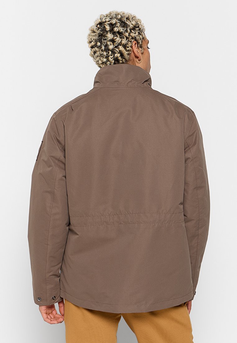 Giacca isolante marrone con colletto alto, tasche con zip e vita leggermente svasata. Tessuto liscio e dettagli minimi.