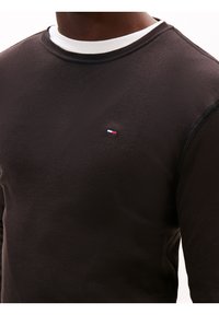 Man wearing a black crewneck sweatshirt with a small blue, white, and red rectangular logo on the chest over a white undershirt.