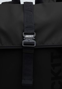 Zaino nero con una texture liscia, caratterizzato da spallacci larghi, una fibbia in plastica e il marchio in rilievo "JACKIT" sulla tracolla.