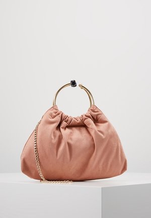 Weiche rosa Wildleder-Handtasche mit gerafftem Oberteil, goldenem Metallringgriff mit schwarzem Akzent und einem goldenen Kettenriemen, der auf einer weißen Oberfläche liegt.