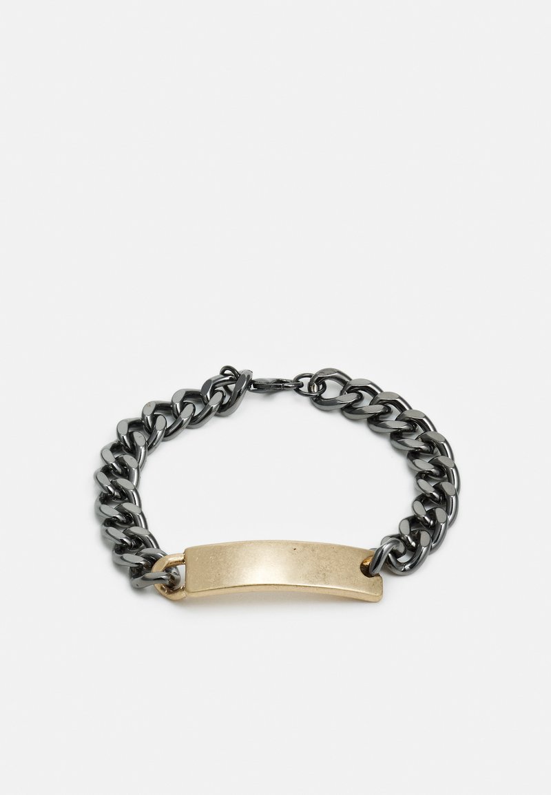 Pulseira de corrente com elos metálicos pretos e uma placa de latão dourada. Apresenta uma textura suave e bordas arredondadas na placa.