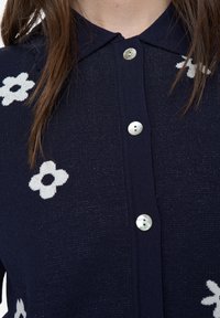 Tmavě modrý pletený cardigan s bílými květinovými vzory, špičatým límcem a kulatými lesklými stříbrnými knoflíky po celé přední straně.