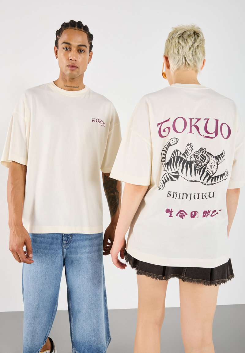 Krém színű, oversized póló, hátul "TOKYO" grafikával és tigris mintával; farmer sorttal viselve. Lazán illeszkedő fazon, rövid ujjú.