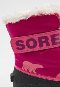 Stivaletto impermeabile rosa con collo in pelliccia sintetica, caratterizzato da una grande etichetta "SOREL" e un design delorso polare in rilievo. Tessuto testurizzato e suola in gomma.