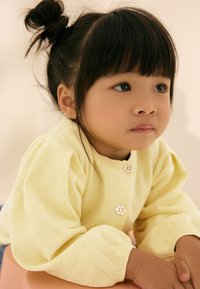 Gul strikket cardigan med puffede ærmer og knapper formet som blomster, båret af et barn med sort hår og lige pandehår. Baggrunden er neutral.