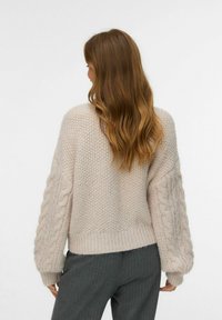 Ljusbeige stickad tröja med strukturerad kropp och flätstickade ärmar, med ribbad nederkant. Bärs över grå kostymbyxor med ränder.