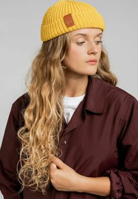 Żółta dziana czapka beanie o prążkowanej teksturze z detalem ze skóry. Noszona z brązową kurtką i białą koszulką pod nią.