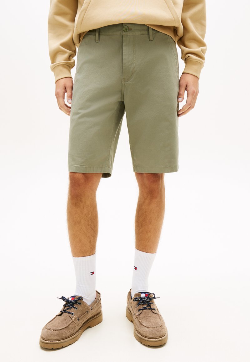 Homme portant une chemise beige à manches longues, un short olive de longueur genou, des chaussettes blanches montantes avec un logo et des chaussures en daim marron clair à lacets.