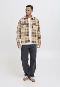 Beige plaid flannel shirt with brown accents and two chest pockets, worn over a white t-shirt, paired with dark grey wide-leg jeans.