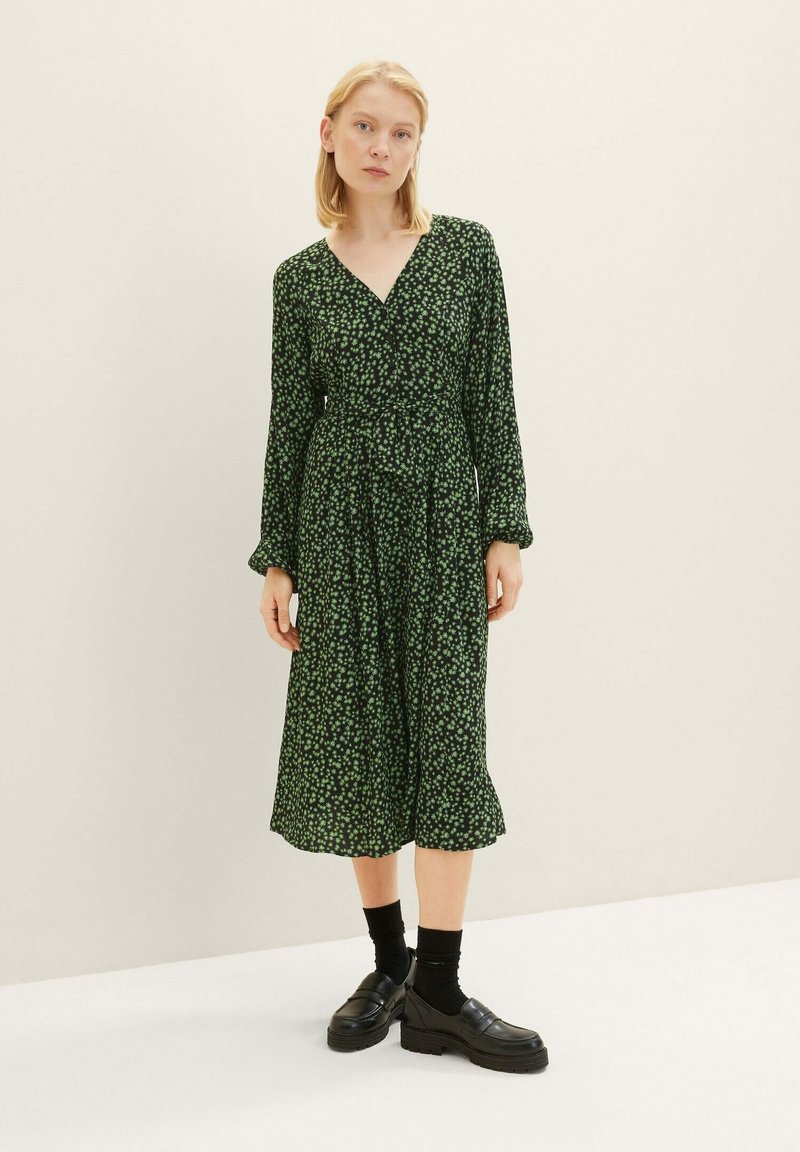 Frau in einem schwarzen Midikleid mit einem kleinen grünen Blumenmuster, langen Ärmeln, schwarzen Socken und klobigen schwarzen Slippern, die vor einem einfarbigen Hintergrund steht.