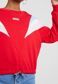 Červená mikina s bílými a světle modrými diagonálními pruhy, žebrovanými manžetami a stahovacím lemem s logem značky na prsou.