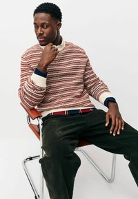 Red and white patterned sweater with a crested neckline, paired with dark green corduroy pants. Subject sits in a chrome chair.