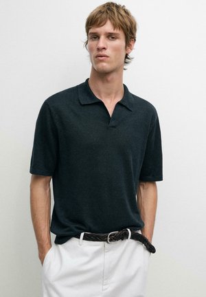 Giovane uomo che indossa una polo nera a maniche corte infilata in pantaloni bianchi con una cintura nera intrecciata, in piedi contro uno sfondo chiaro e uniforme.