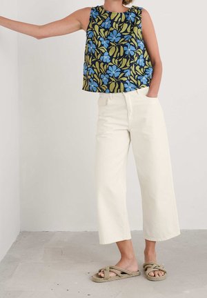 Kobieta ubrana w kwiecistą, bezrękawnikową bluzkę z niebieskimi i zielonymi liśćmi, kremowe szerokie spodnie o długości do połowy łydki oraz sandały z sznurka, stojąca w pomieszczeniu.