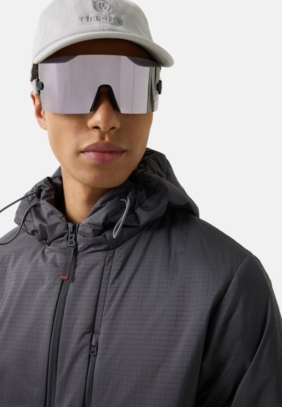 Jong persoon met grote, spiegelende vizierzonnebril, witte pet en donkergrijze capuchonjas met rits en trekkoord.