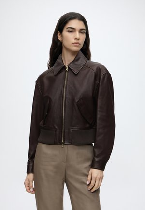 Frau mit langen dunklen Haaren trägt eine dunkelbraune Lederjacke mit Reißverschluss und beige Hose, steht vor einem schlichten hellen Hintergrund.