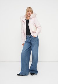Světle růžová péřová bunda s kapucí lemovanou kožešinou, v kombinaci s modrými džíny širokého střihu. Modelka drží malou, světle barevnou kabelku.