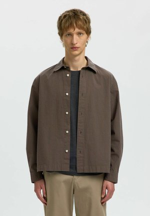 Ung mand med kort lyst brunt hår iført en brun ternet skjorte over en mørkegrå t-shirt og beige bukser, stående mod en hvid baggrund.
