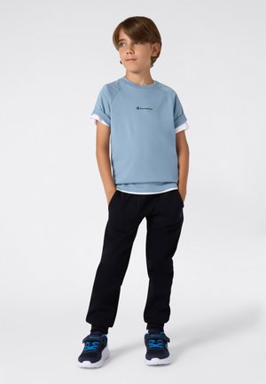 Jongen met bruin haar in een lichtblauw Champion T-shirt, zwarte broek en zwart-blauwe sneakers, staande met zijn handen in zijn zakken.