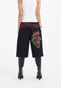 Mustat farkkushortsit, joissa on brodeerattu tiikeri- ja punainen sydänkuvio, ja jotka on koristeltu strasseilla. Vyötärönauhassa on punaisella brodeerattu 'Ed Hardy' -teksti.