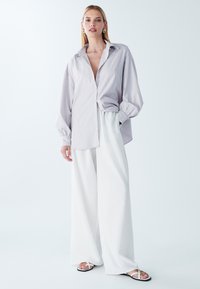 Gestreifte Button-Down-Bluse in hellem Lavendel, lässige Passform mit Ballonärmel. Kombiniert mit weit geschnittenen weißen Hosen und flachen Sandalen.