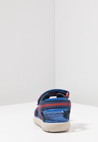 Blå och grå sandal med röda accenter, utrustad med justerbara remmar, texturerad innersula och en flexibel sula designad för komfort och stöd.