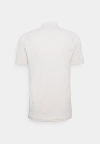 Ljus beige polo med hög krage, korta ärmar och en mjuk textur; har en subtil logotypdetalj och vita accenter på sidan.