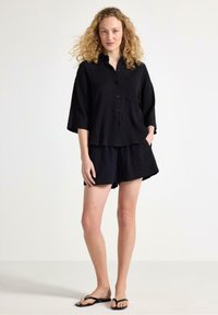 Svart linne skjorta med kort ärm och matchande shorts, med knäppning fram och avslappnad passform. Bärs med minimalistiska svarta flip-flops.