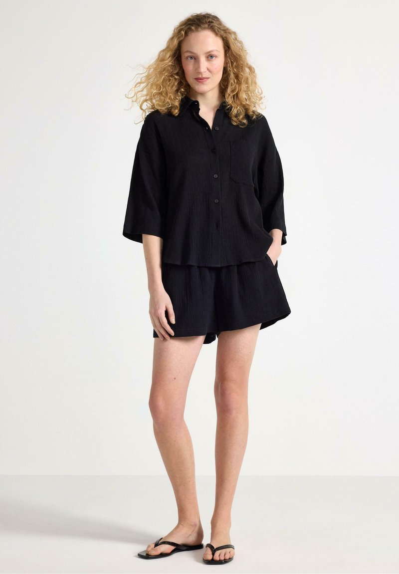 Svart linne skjorta med kort ärm och matchande shorts, med knäppning fram och avslappnad passform. Bärs med minimalistiska svarta flip-flops.