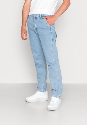 Man wearing light blue straight-leg jeans with side loop detail, white sneakers, and a white short-sleeve shirt, standing with one hand in pocket.