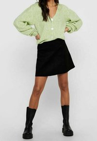 Hellgrüne gestrickte Strickjacke mit übergroßen Ärmeln und weißen Knöpfen, kombiniert mit einem schwarzen Minirock und kniehohen schwarzen Stiefeln.