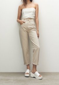 Femme portant un haut blanc sans bretelles, un pantalon beige taille haute avec revers, et des sandales blanches à plateforme, debout contre un mur neutre.