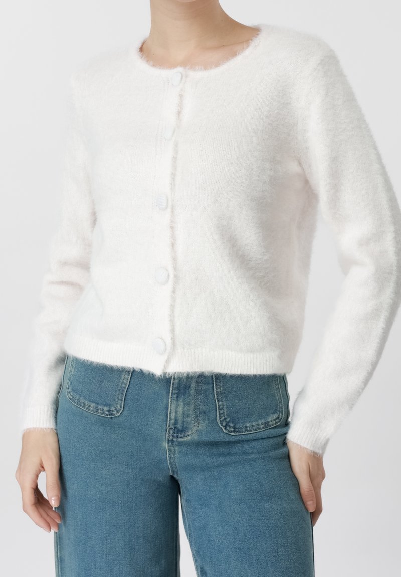 Femme portant un cardigan blanc doux et duveteux boutonné avec des manches longues et un jean bleu taille haute sur un fond uni.