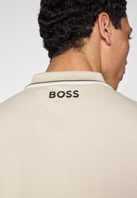 Bézs színű pólóing fekete-fehér gallérral, a hátoldalon fekete "BOSS" hímzéssel. A szövet texturált felületű.