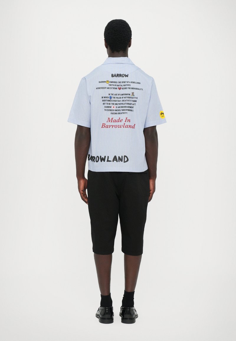 Camicia a righe blu e bianche con maniche corte che presenta grafiche testuali sulla parte posteriore, abbinata a pantaloni corti neri fino al ginocchio e scarpe nere lucide.
