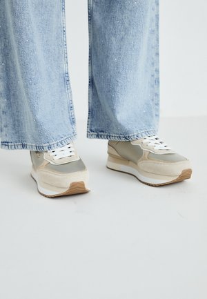 Bežīgi un pelēki sporta apavi ar samtainu un atstarojošu materiālu kombināciju, baltām šņorēm, gumijas zoli un teksturētām akcentiem. Nēsāti ar gaiši ziliem džinsu biksēm.