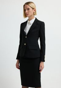 Blazer nero su misura con bottoni dorati, due tasche frontali e una forma aderente. Indossato sopra una camicia bianca, abbinato a una gonna coordinata.