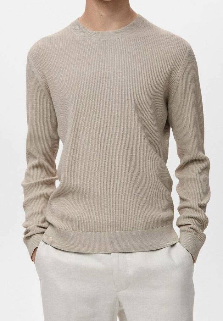 Homme portant un pull beige côtelé à manches longues et un pantalon de couleur claire, les mains dans les poches, devant un fond uni.