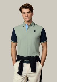 Joven con camiseta tipo polo verde y azul marino, pantalones beige y un suéter negro atado a la cintura, de pie con las manos en los bolsillos.