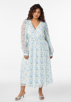Kobieta ubrana w długą sukienkę midi w kolorach jasnoniebieskim i białym z kwiatowym wzorem, z dekoltem w serek i marszczonym pasem, stojąca na białym tle.