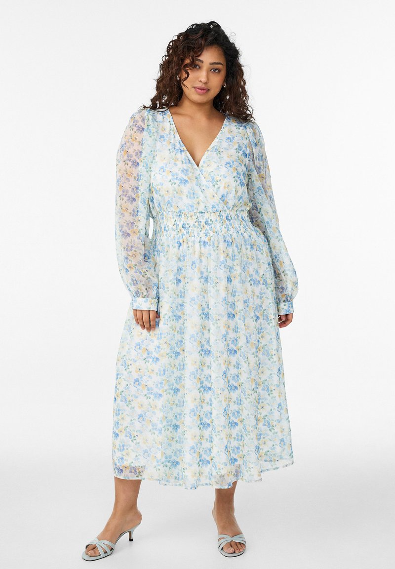 Frau trägt ein langärmliges, hellblau-weißes Midikleid mit Blumenmuster, V-Ausschnitt und geraffter Taille, steht vor einem weißen Hintergrund.