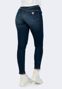 Jeans skinnies azul oscuro con una cintura media, bolsillos traseros y un logo en forma de triángulo. Llevados con zapatillas blancas que cuentan con una suela texturizada.