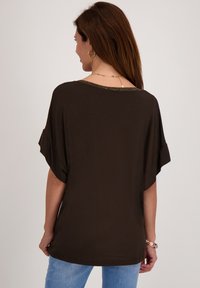 Bruine, losse top met korte mouwen en een ronde halslijn. Heeft een decoratieve metalen afwerking langs de halslijn.