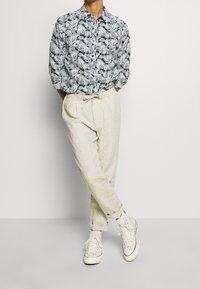 Persona con una camicia a fantasia di foglie bianco e nero, pantaloni beige con cordoncino e sneakers bianche alte, in piedi con le gambe incrociate e le mani nelle tasche.